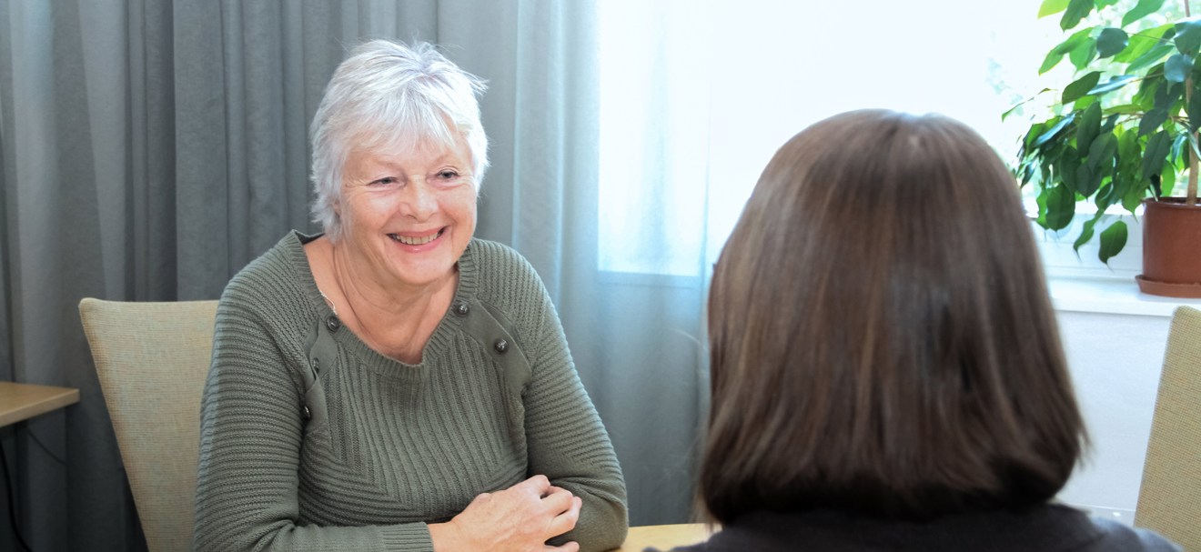 Leende kvinna med kort, vitt hår tittar på en person på andra sidan bordet, vars bakhuvud syns.  