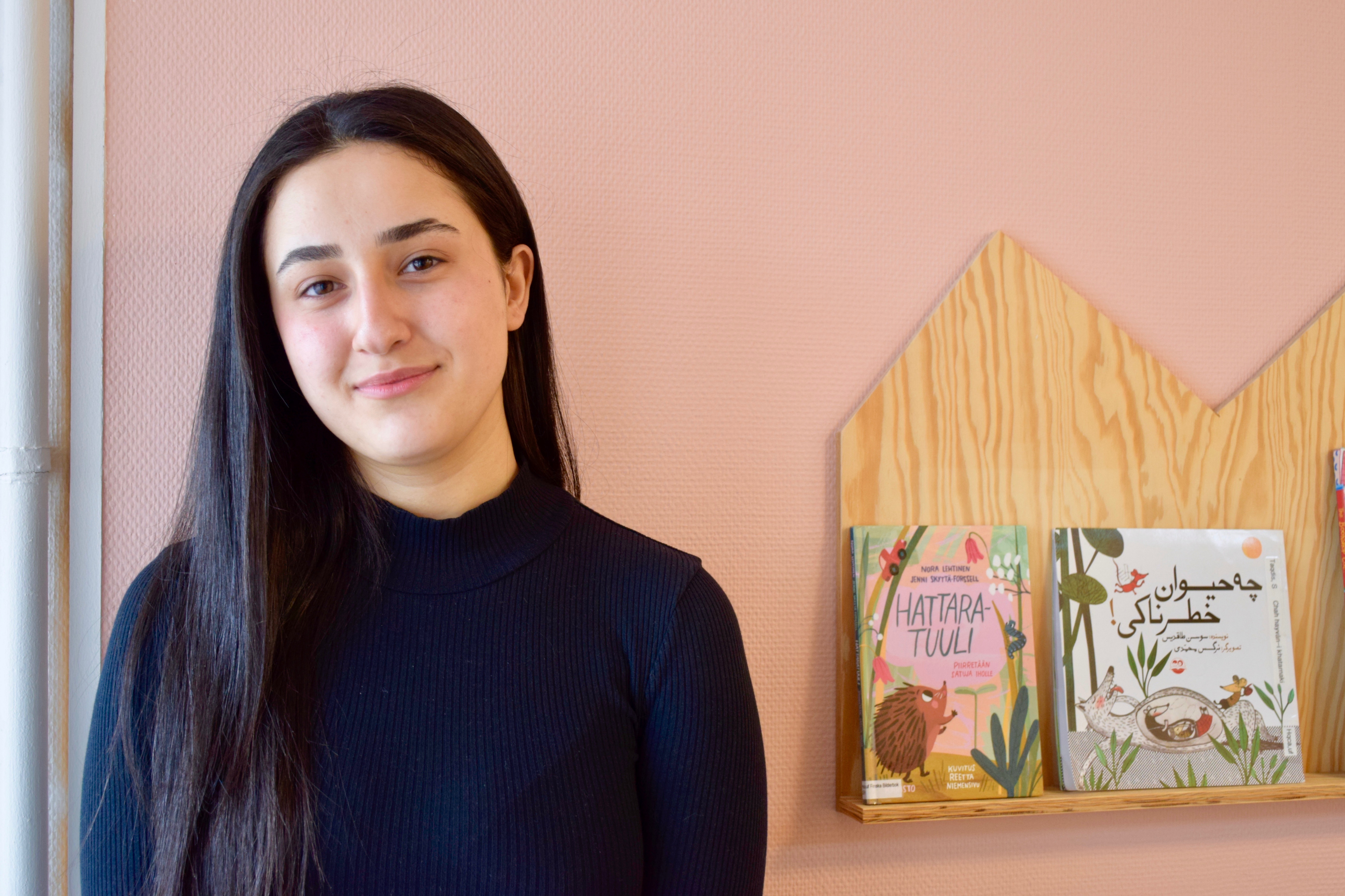 Shana Sulaiman har långt mörkt hår och är klädd i en svart tröja. Bakom henne finns en rosamålad vägg och på väggen sitter en bokhylla med böcker. 