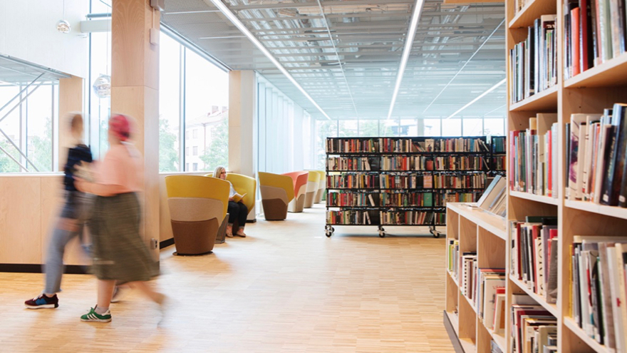 Örebro stadsbibliotek. Stora glasväggar ger stort insläpp, längs glasväggen står färgglada fåtöljer på rad. Ett par kvinnor passerar fotografen, fyllda bokhyllor står både till höger och mitt fram.