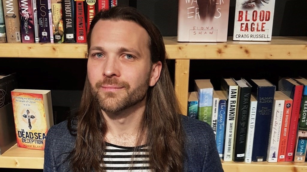 Pontus Kjellberg har långt brunt hår och skägg. Bakom honom finns en bokhylla fylld med böcker. 