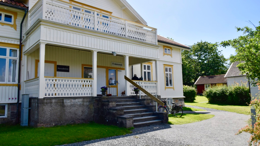 En vit äldra byggnad i trä. Huset har en stor veranda där biblioteket har sin entré. Solen skiner och huset är omgivet av en trädgård. 