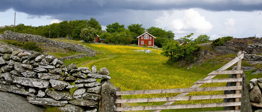 Ett rött hus med vita detaljer står på en äng. En stengärsgård omgärdar huset och en trägrind delar gärsgården.