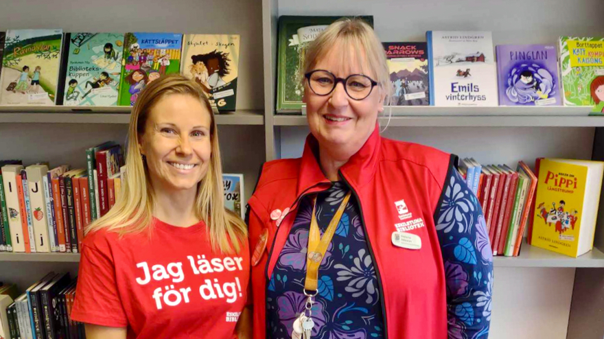 Annika har blont långt hår och är klädd i en röd t-shirt med texten "Jag läser för dig!". Helena har blont uppsatt hår, bär glasögon och bär en röd väst ovanpå en mönstrad jumper. De står nära varandra och ler mot kameran, i bakgrunden finns en bokhylla.  