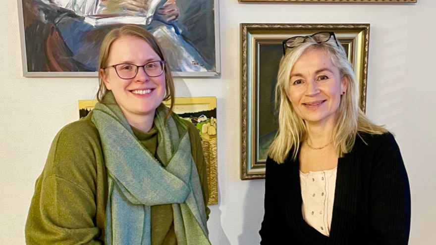 Raisa Leussu och Susanne Redebo sitter framför en vägg med tavlor. Susanne sitter till höger, hon har blont långt hår, en vit blus och en svart kofta. Raisa sitter till vänster och bär en grön tröja och en grön scarf, hon har blont uppsatt hår.  